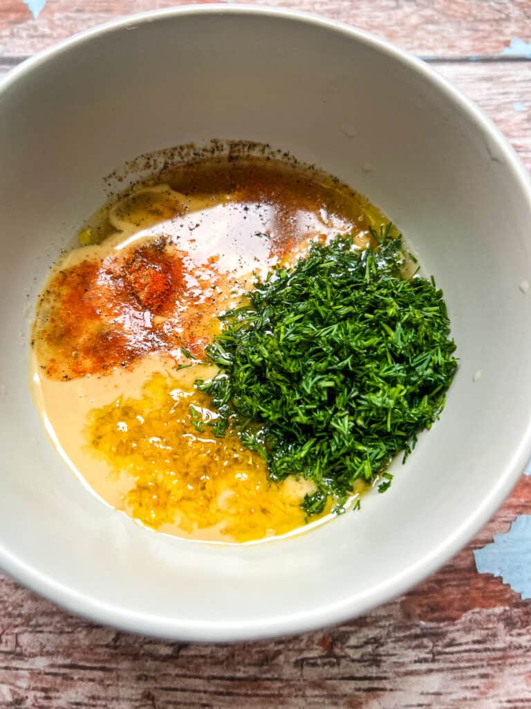 white bowl with tahini, lemon zest, lemon juice, red cayenne, cumin, olive oil, vinegar, and chopped fresh dill