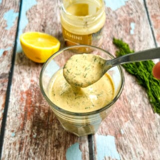glass with mixed tahini dressing in the foreground with a spoon poring the dressing, showing the thick and creamy texture, half a lemon, jar of tahini, and fresh dill in the background