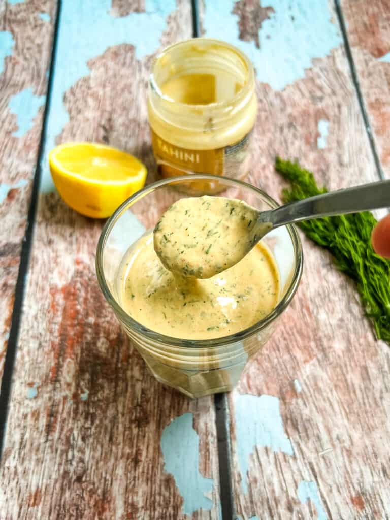 glass with mixed tahini dressing in the foreground with a spoon poring the dressing, showing the thick and creamy texture, half a lemon, jar of tahini, and fresh dill in the background