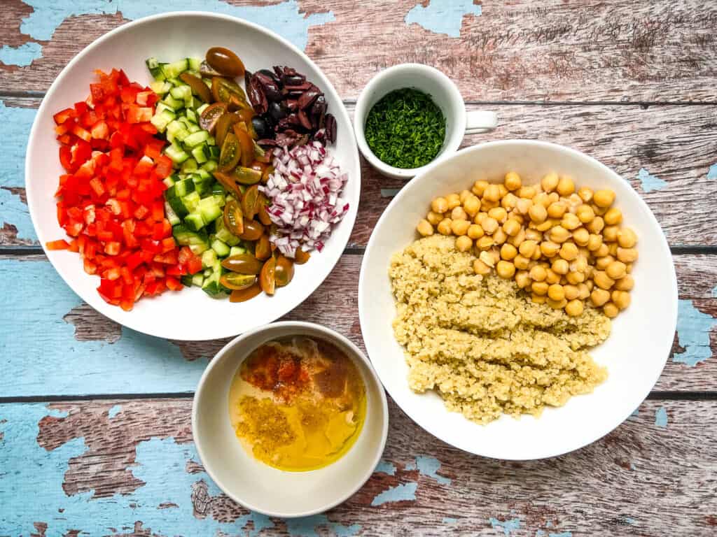 prepped ingredients arranged, chopped red bell pepper, cucumber, tomatoes, red onion, black olives, cup with minced dill, bowl with chickpeas and quinoa, bowl of tahini dressing ingredients, unmixed