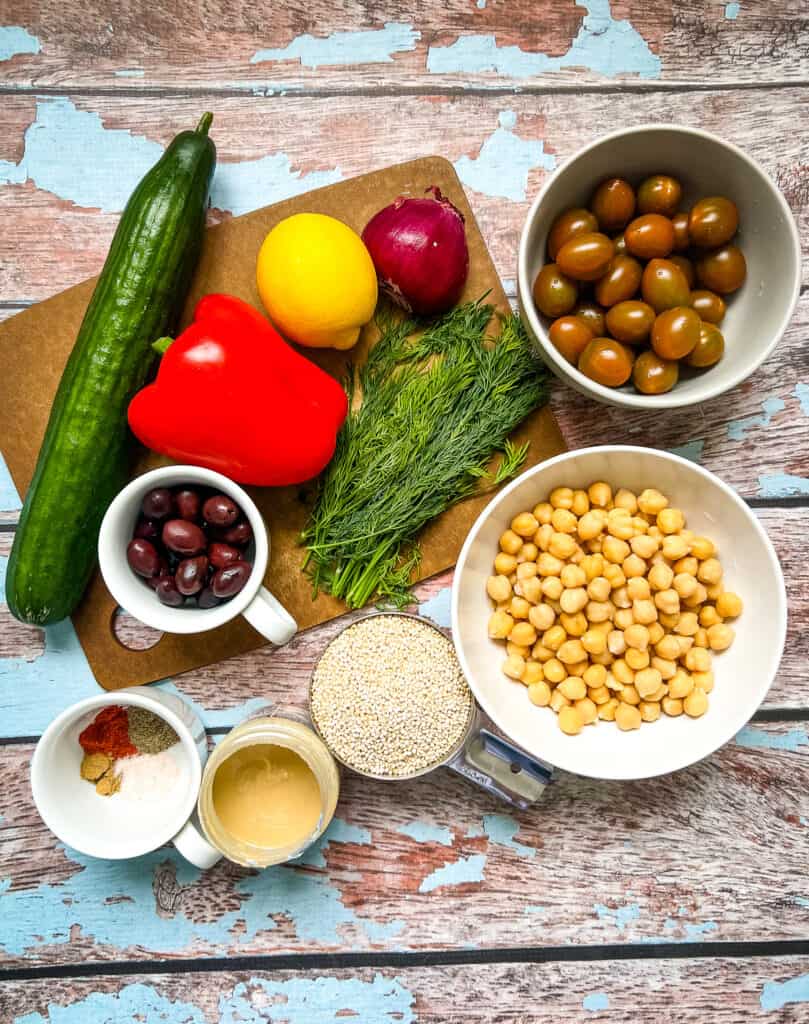 displayed ingredients, whole english cucumber, red bell pepper, lemon, red onion, grape tomatoes, chickpeas in a bowl, uncooked quinoa, tahini, spices in a cup, olives in a cup, fresh dill