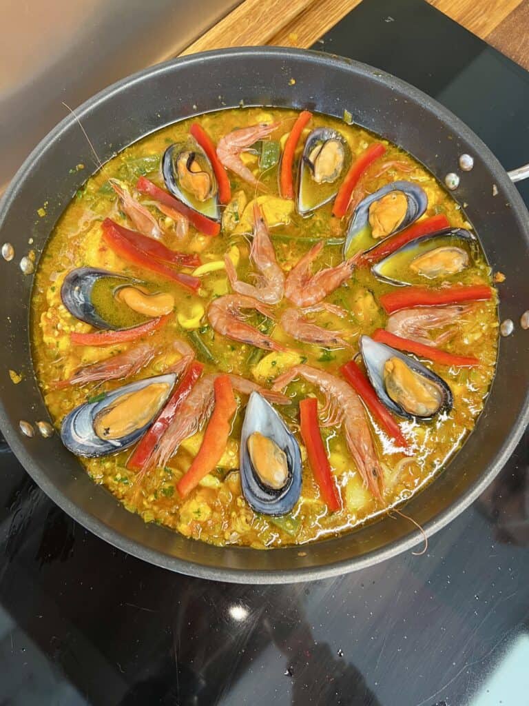 large paella pan with rice, broth, chicken, prawns, mussels, green beans, and sliced red peppers fanned out decoratively.