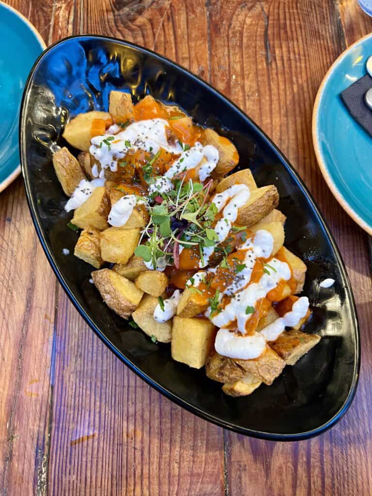 black dish of crispy fried potatoes with red sauce, white sauce, and topped with green herbs