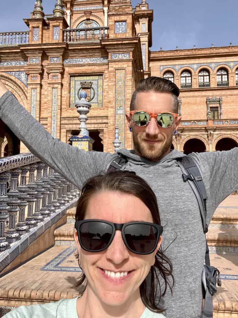 two adults, one male, one female, in sunglasses, smiling in front of the main building at plaza de espana