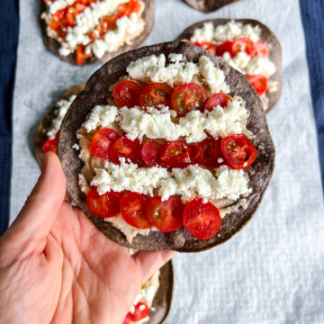 Hand holding one blue corn tostadas are topped with white bean puree, with salsa, tomatoes, and crumbly cheese arranged to resemble the red and white stripes of the USA flag.