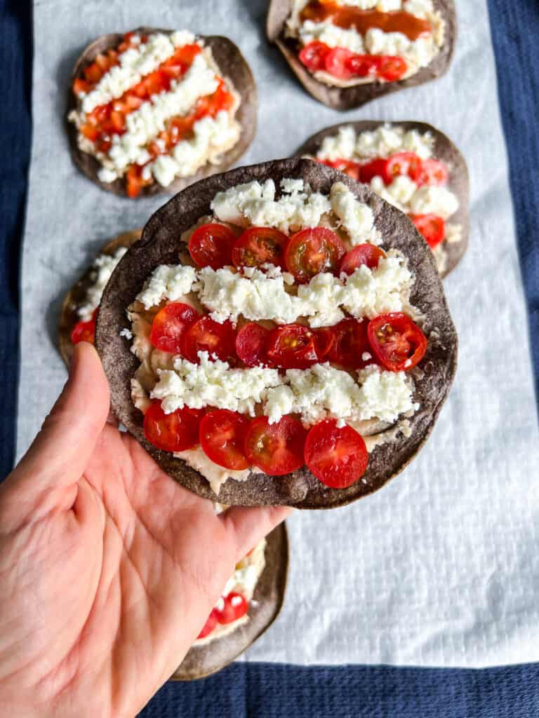 Hand holding one blue corn tostadas are topped with white bean puree, with salsa, tomatoes, and crumbly cheese arranged to resemble the red and white stripes of the USA flag. 