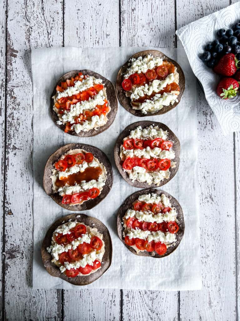 Blue corn tostadas are topped with white bean puree, with salsa, tomatoes, and crumbly cheese arranged to resemble the red and white stripes of the USA flag with a plate of blueberries and strawberries in the corner.