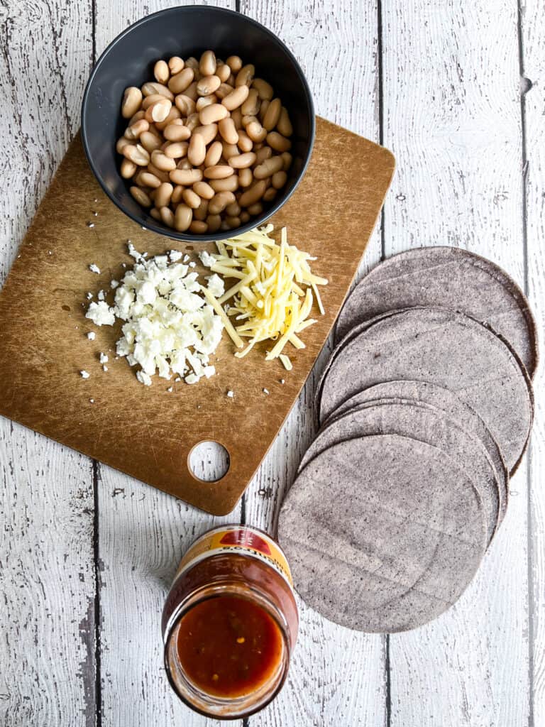 Cutting board displaying ingredients: crumbled white cheese and shredded cheddar, bowl of white beans, jar of salsa, and 6 blue corn tortillas.