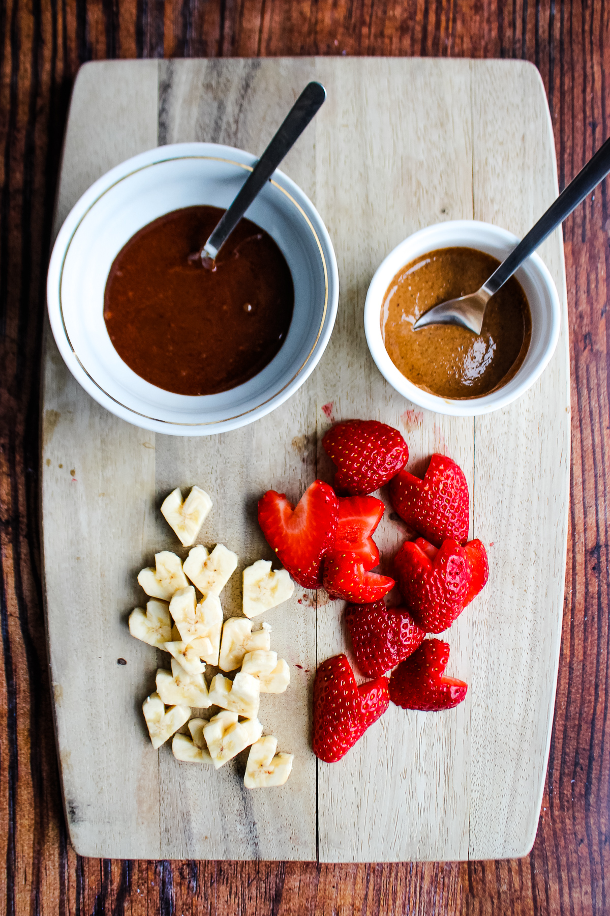 Heart shaped bananas, heart shaped strawberries on cutting board, a small ramekin of chocolate spread and a small ramekin of nut butter.