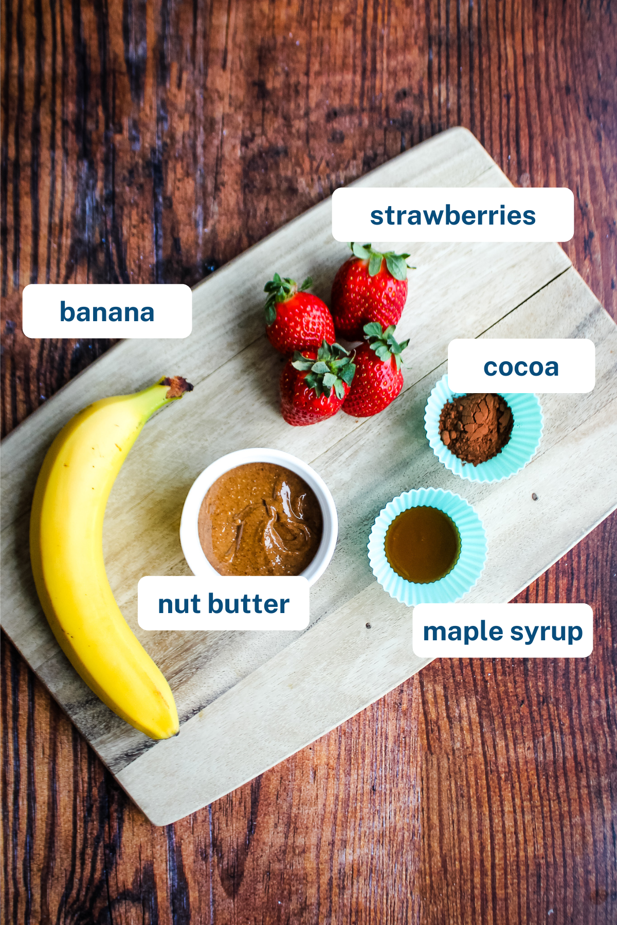 A banana, 4 strawberries, cocoa powder, maple syupr, and almond butter displayed on a cutting board with labels.