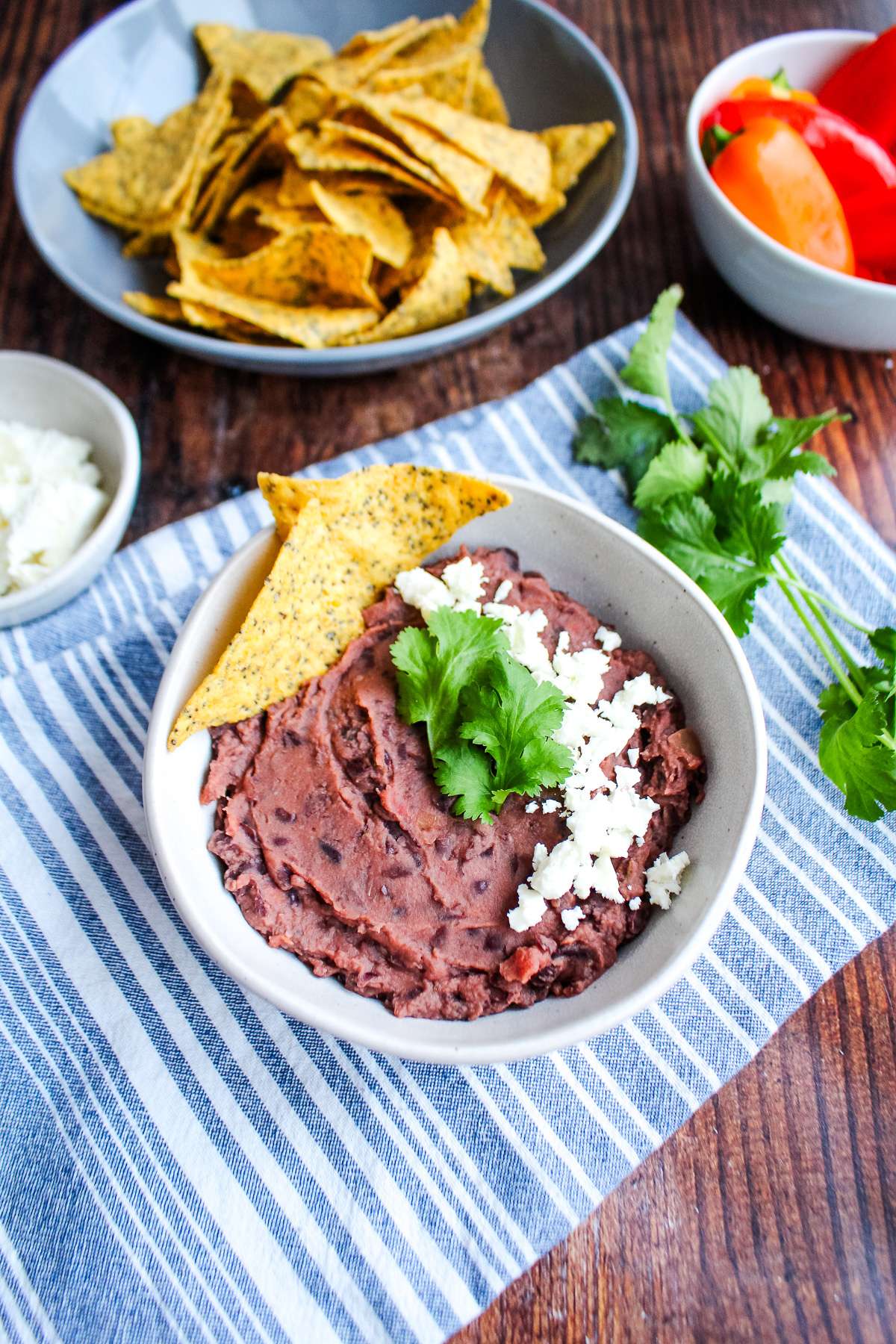 A bowl of easy black bean dip on the table with a bowl of chips and chilies in the background.