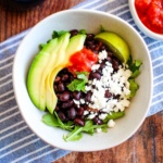 A bowl of black bean avocado salad on a blue tea towel with sliced avocado and cheese crumbles.