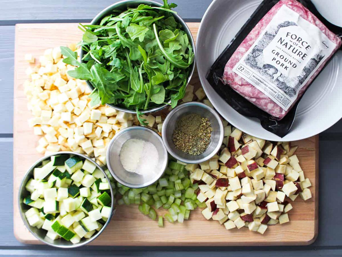 cutting board displaying diced root vegetables, arugula, spices, salt, and ground pork