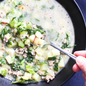 A bowl of ground pork soup with vegetables on the table with a hand easing a soup into the bowl and lifting up a bite.