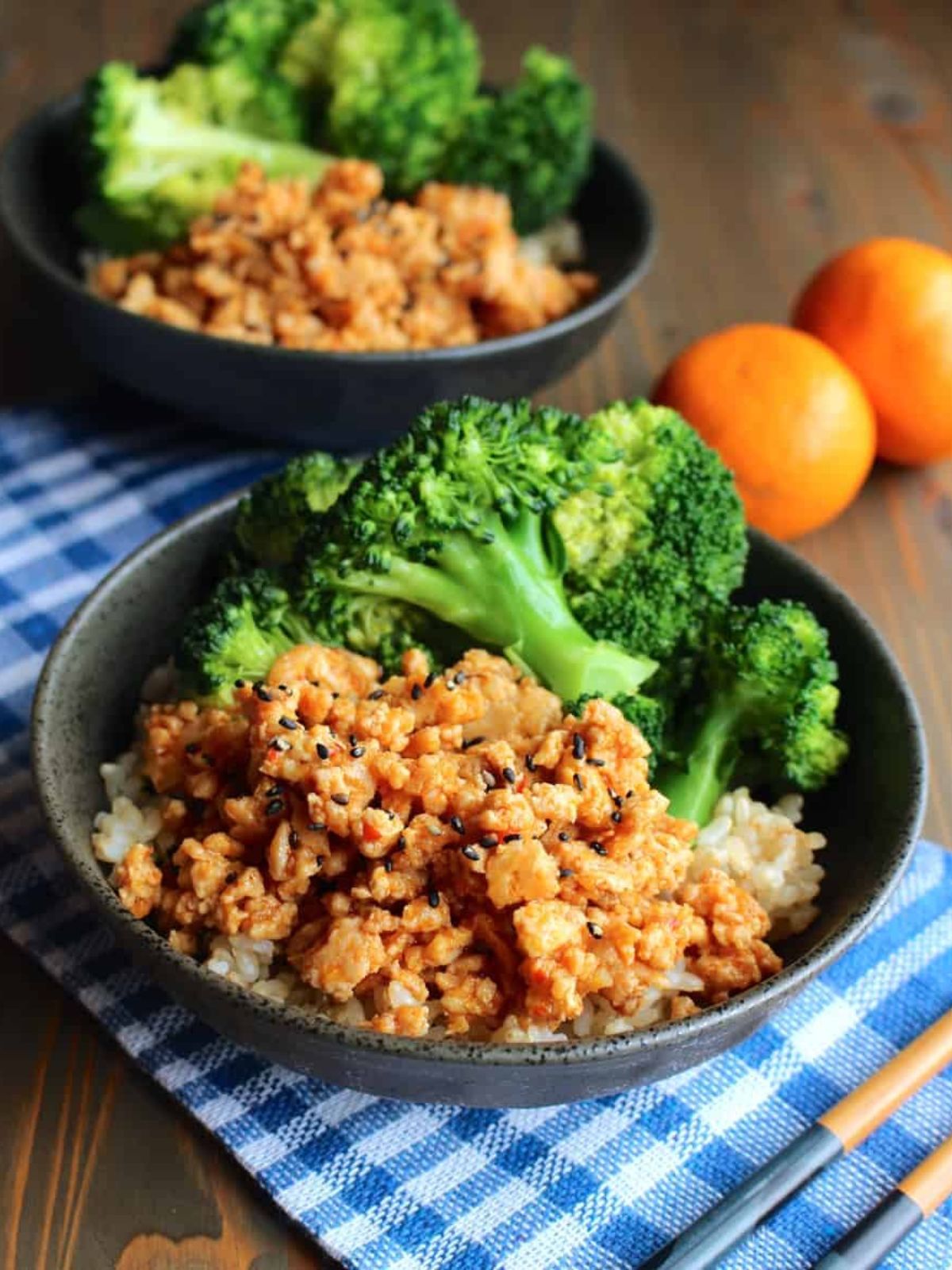 A bowl of a ground chicken rice bowl with broccoli on the table with a blue and white checkered towel and fresh oranges.