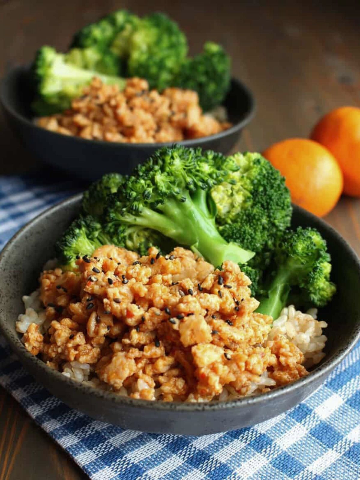 A orange ground chicken rice bowl on the table with mandarins to the side and an extra bowl in the back.