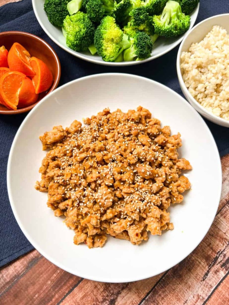 White bowl with orange ground chicken and rice, broccoli, and orange slices in the background.