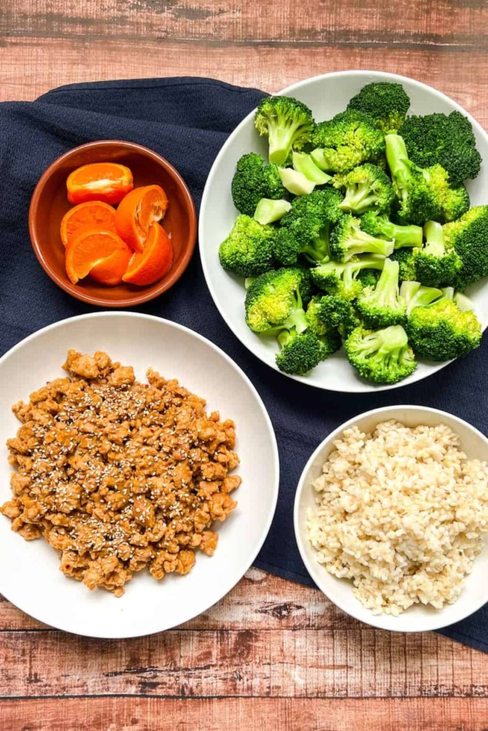 Display of components of this meal, with ground orange chicken, rice, broccoli, and orange wedges in different bowls.