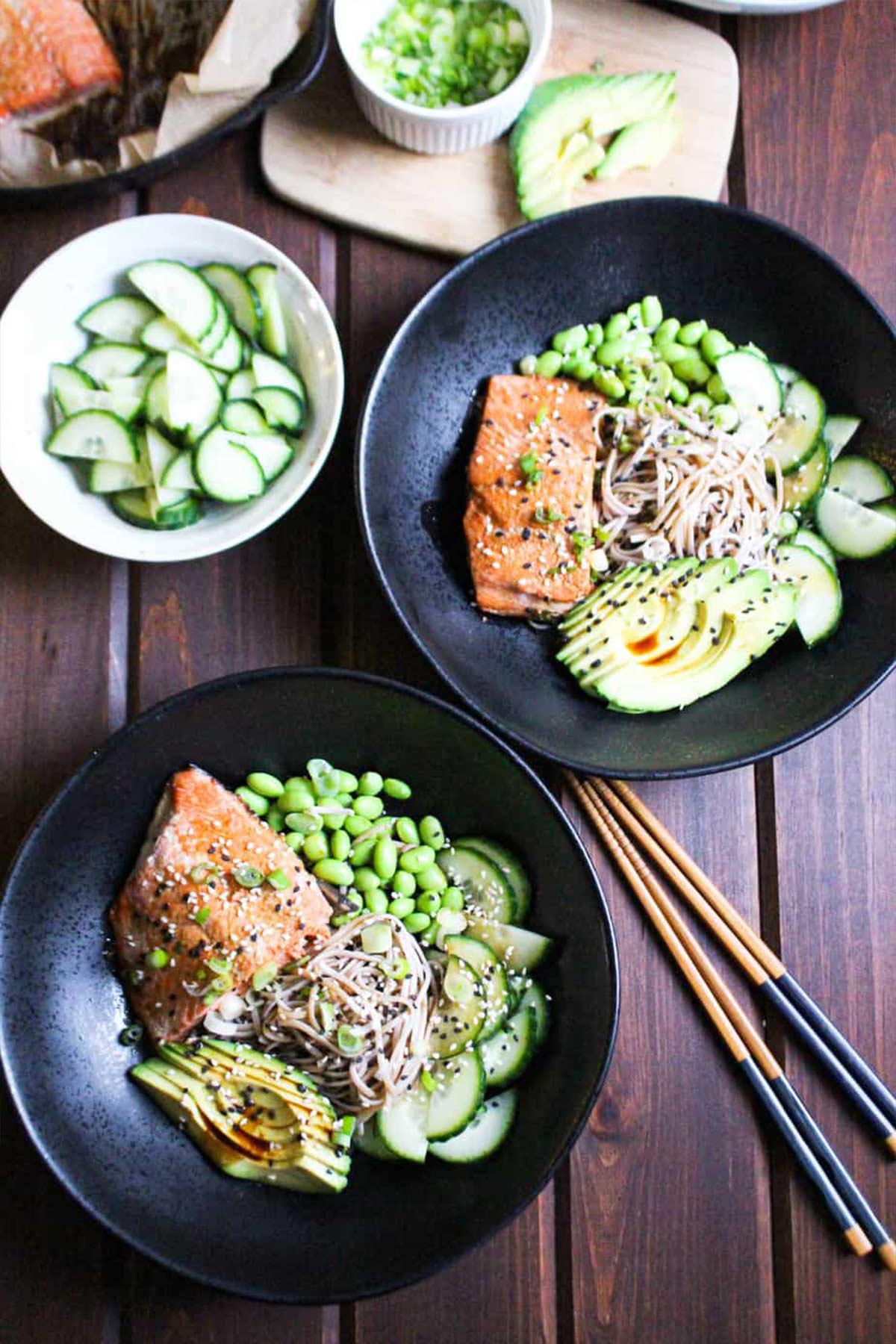 Two salmon noodle bowls on the table each with salmon, edamame, cucumber slices, avocado slices, buckwheat soba, and sauce