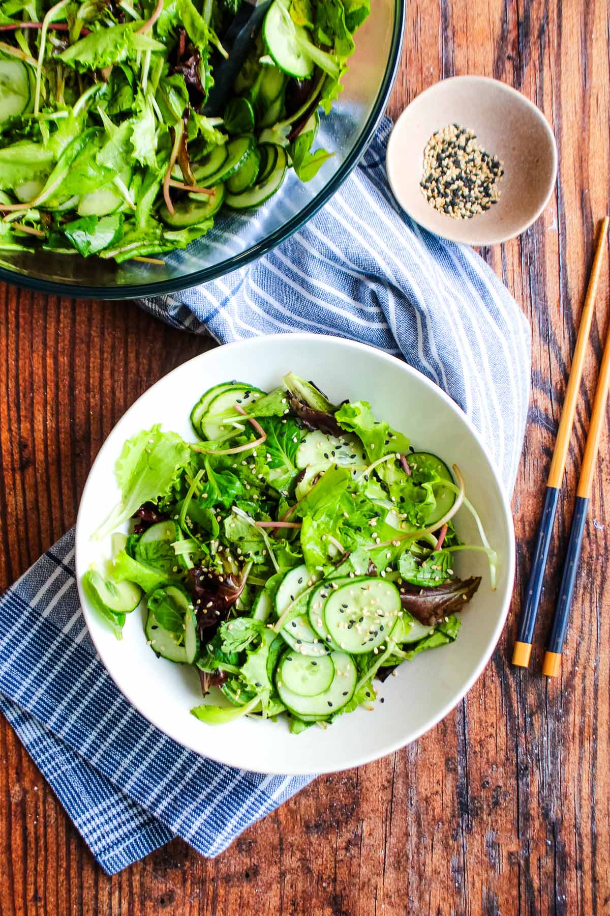 Sesame cucumber salad on the table with a blue tea towel, chopsticks, a bowl of greens, and sesame seeds.