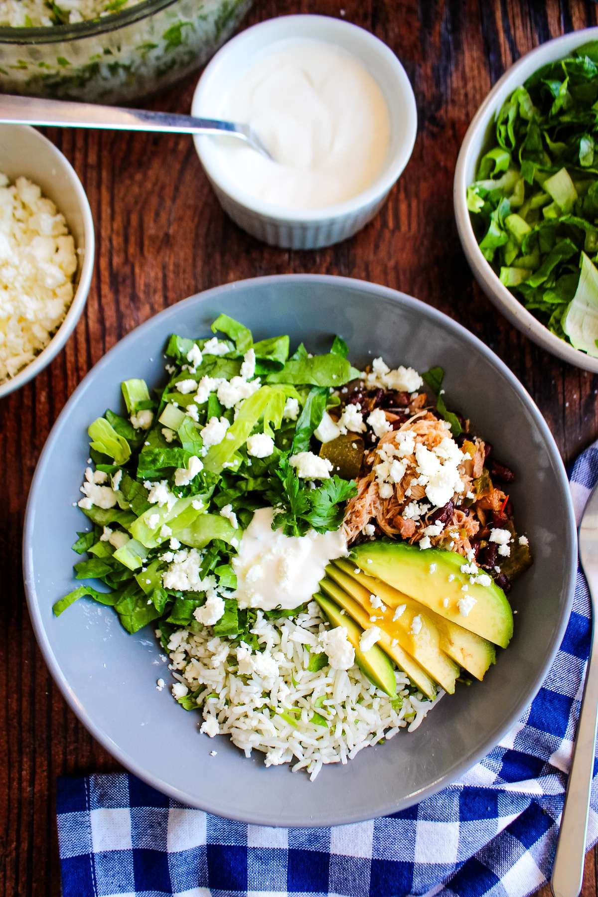 A slow cooker burrito bowl on the table with rice, avocado, lettuce and shredded chicken.