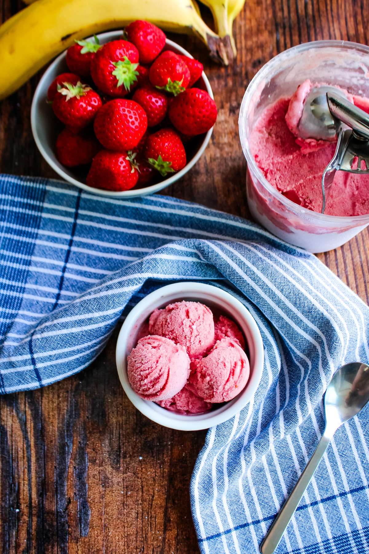 A bowl of strawberry and banana ice cream on the table with a bowl of strawberries and a container of more ice cream with a scoop in the corner.