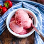 A bowl of strawberry banana ice cream with a spoon on a table with a blue napkin and fresh berries.