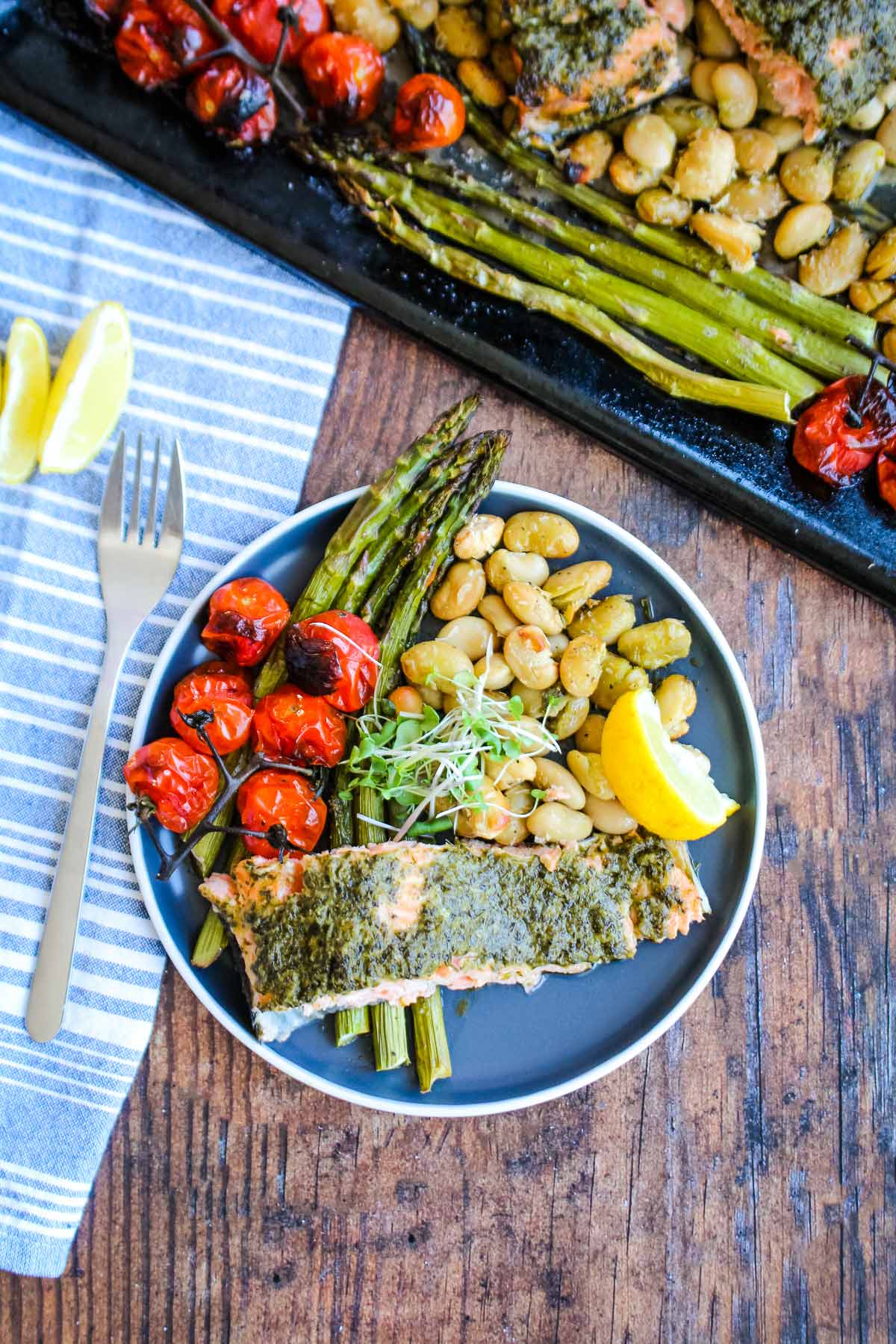 Pesto salmon served on a blue plate with veggies on the side and more on the sheet pan in the background.