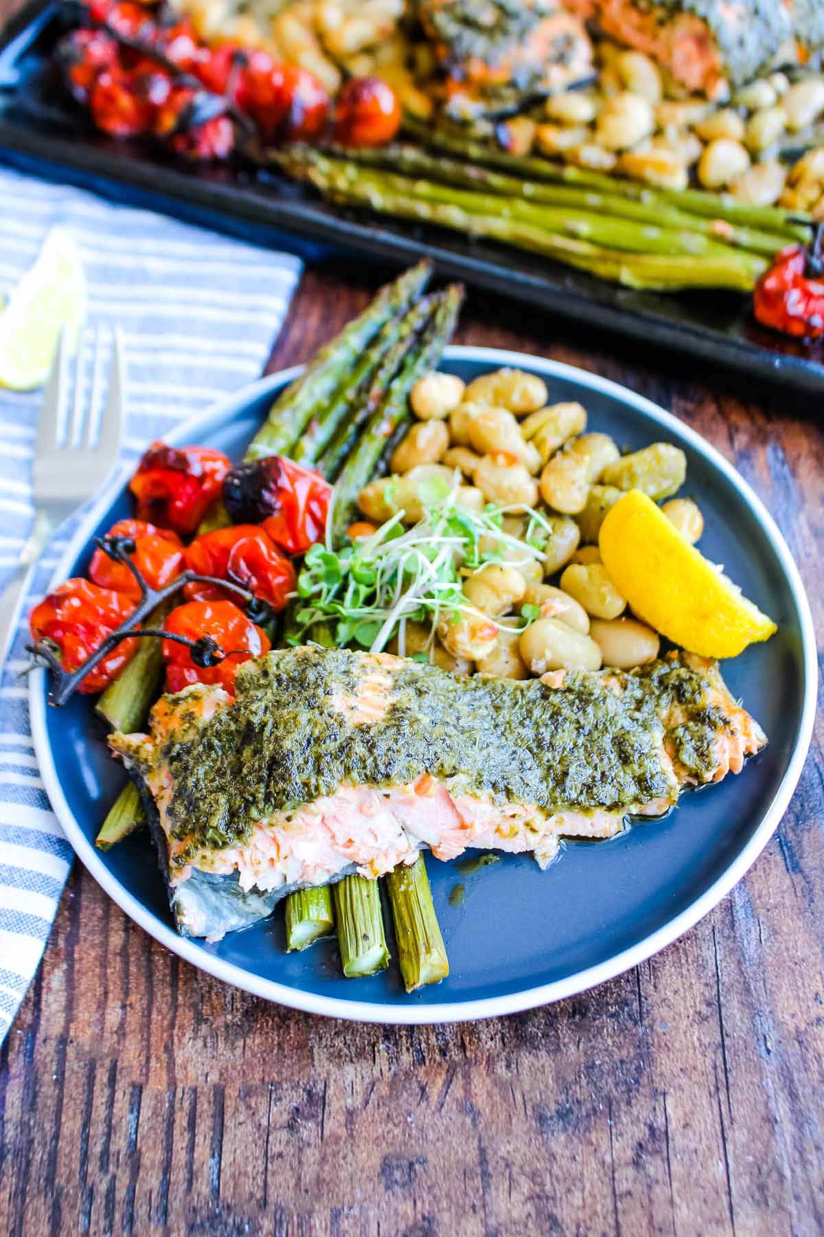 Pesto salmon and vegetables on a plate with the sheet pan of veggies in the backgorund.