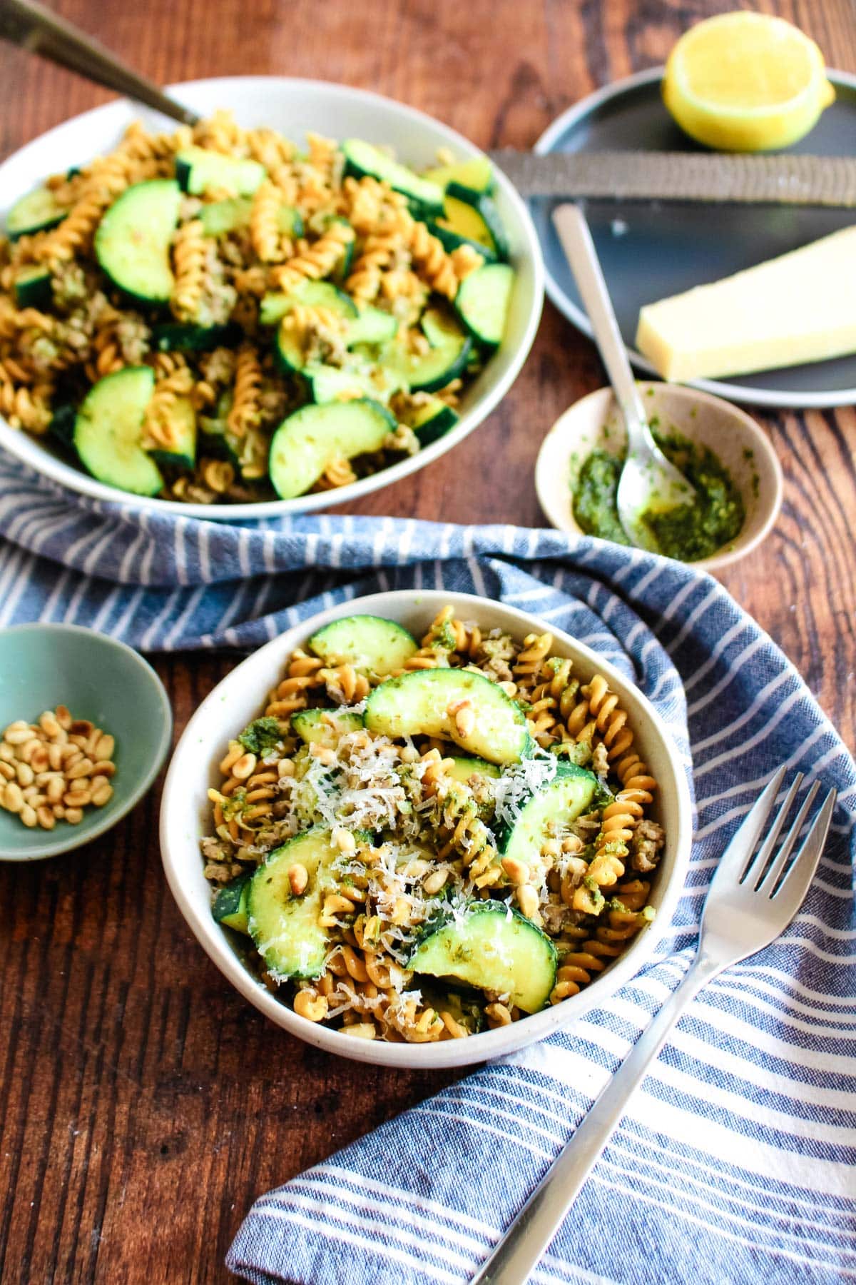 Two bowls of zucchini pesto pasta served up on the table with little bowls of pine nuts and pesto.