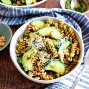 Zucchini pesto pasta served up in a white bowl on the table with blue tea towel and small condiment bowls in the background.