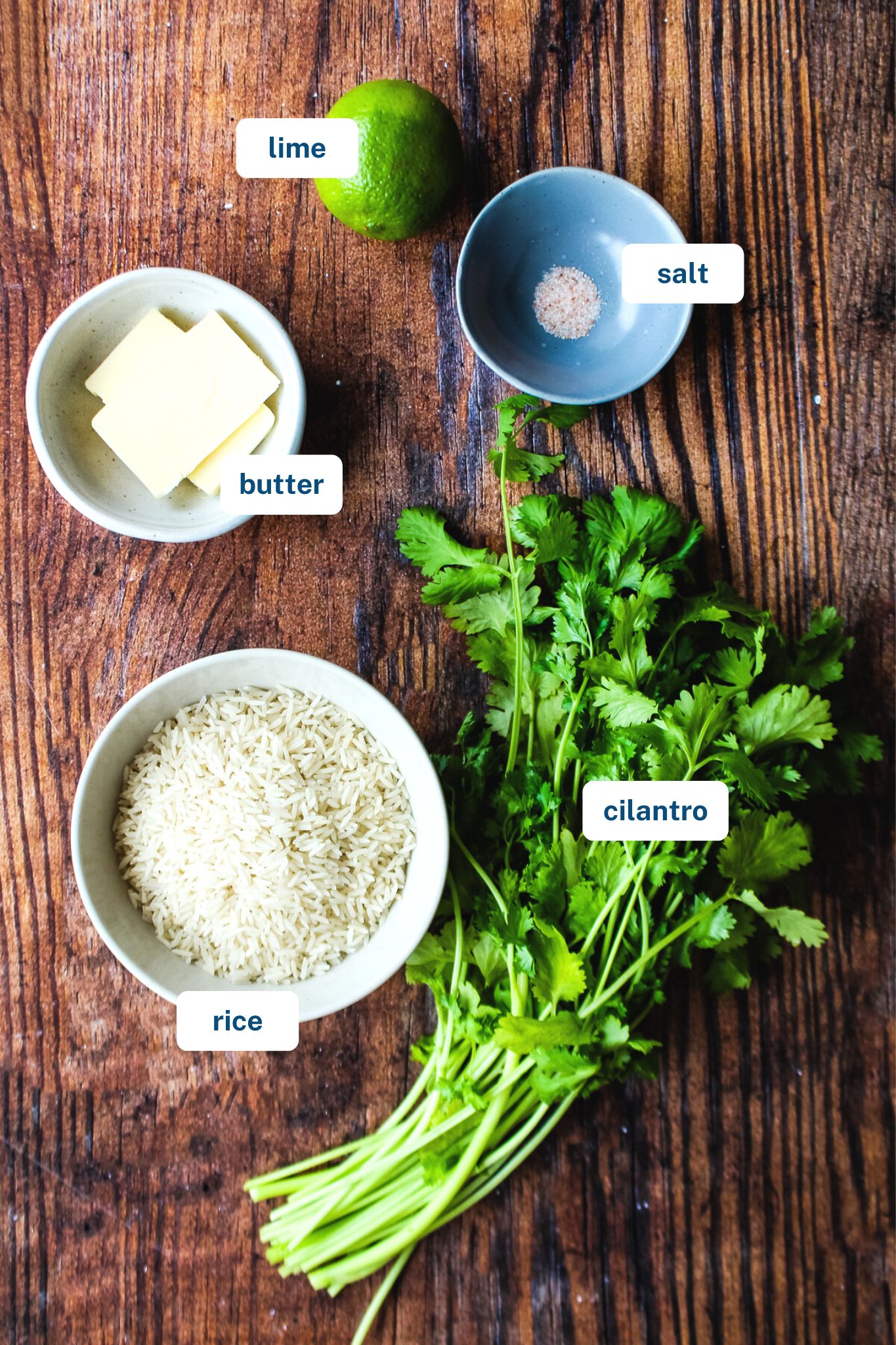 brown wooden background displaying lime, salt, fresh cilantro, uncooked white rice, and butter