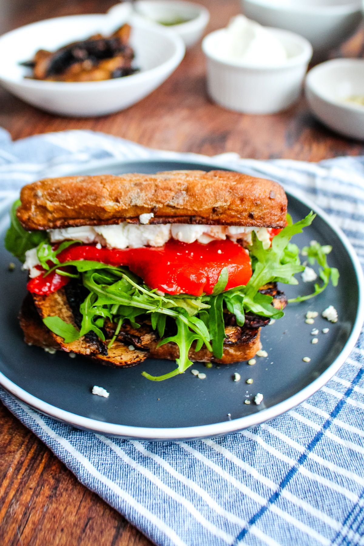 A portobello mushroom sandwich on a blue plate with bowls of ingredients in the background.