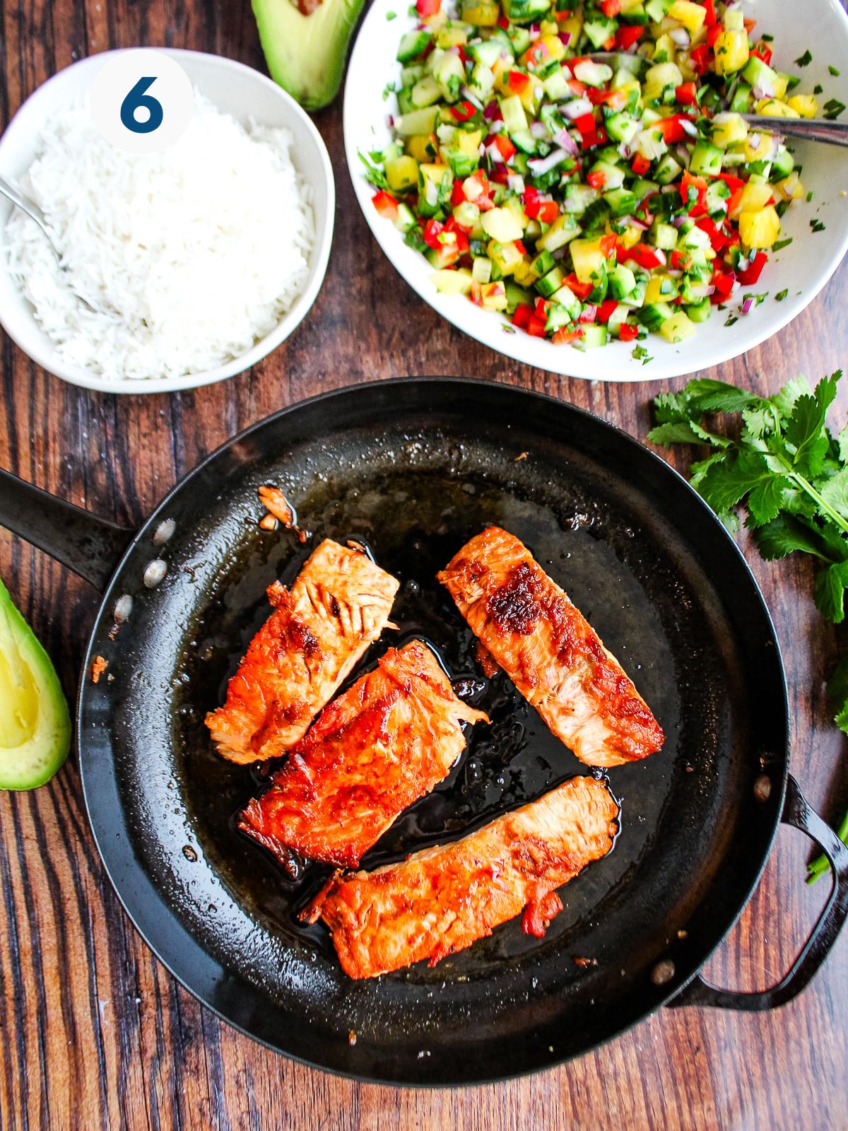 Salmon in the skillet on the table with a bowl of rice and pineapple salsa.