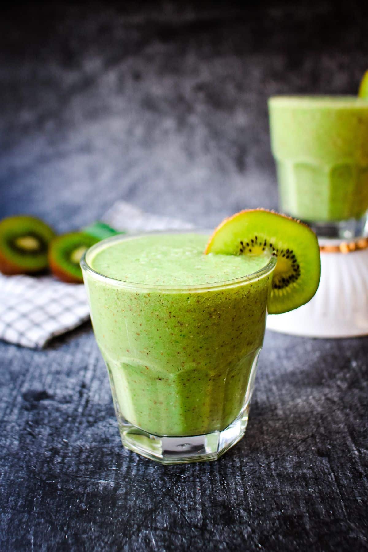 A kiwi smoothie on the table with one in the background to the right.