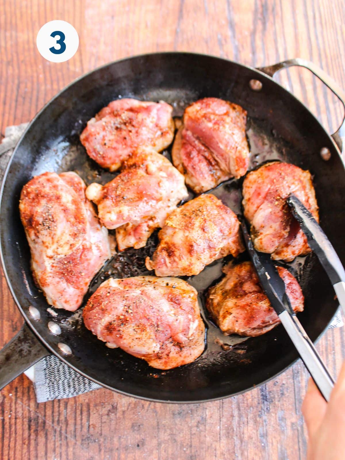 Tongs reaching in to grab a chicken thigh cooking in a cast iron skillet.