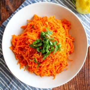 Moroccan carrot salad in a white bowl with a lemon next to the bowl and fresh herbs on top.