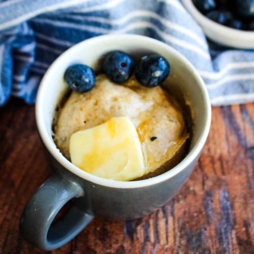 Gluten-free pancake in a mug on a wooden table topped with butter and blueberries.