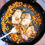 A skillet with chicken and crispy chickpeas and a spoon resting on the edge of the dish.