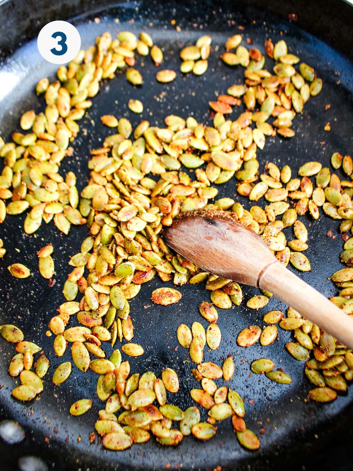 A spoon stirring spiced pumpkin seeds in a skillet.