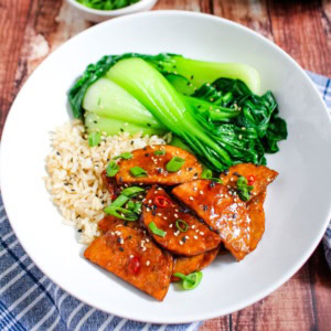 Teriyaki tempeh on a white plate with some brown rice and bok choy.