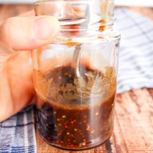 A jar of mustard balsamic dressing held by a hand over the table.