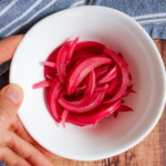 A bowl of pickled red onions on a wooden table with a hand grasping the edge of the bowl.