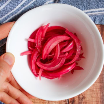 A bowl of pickled red onions on a wooden table with a hand grasping the edge of the bowl.