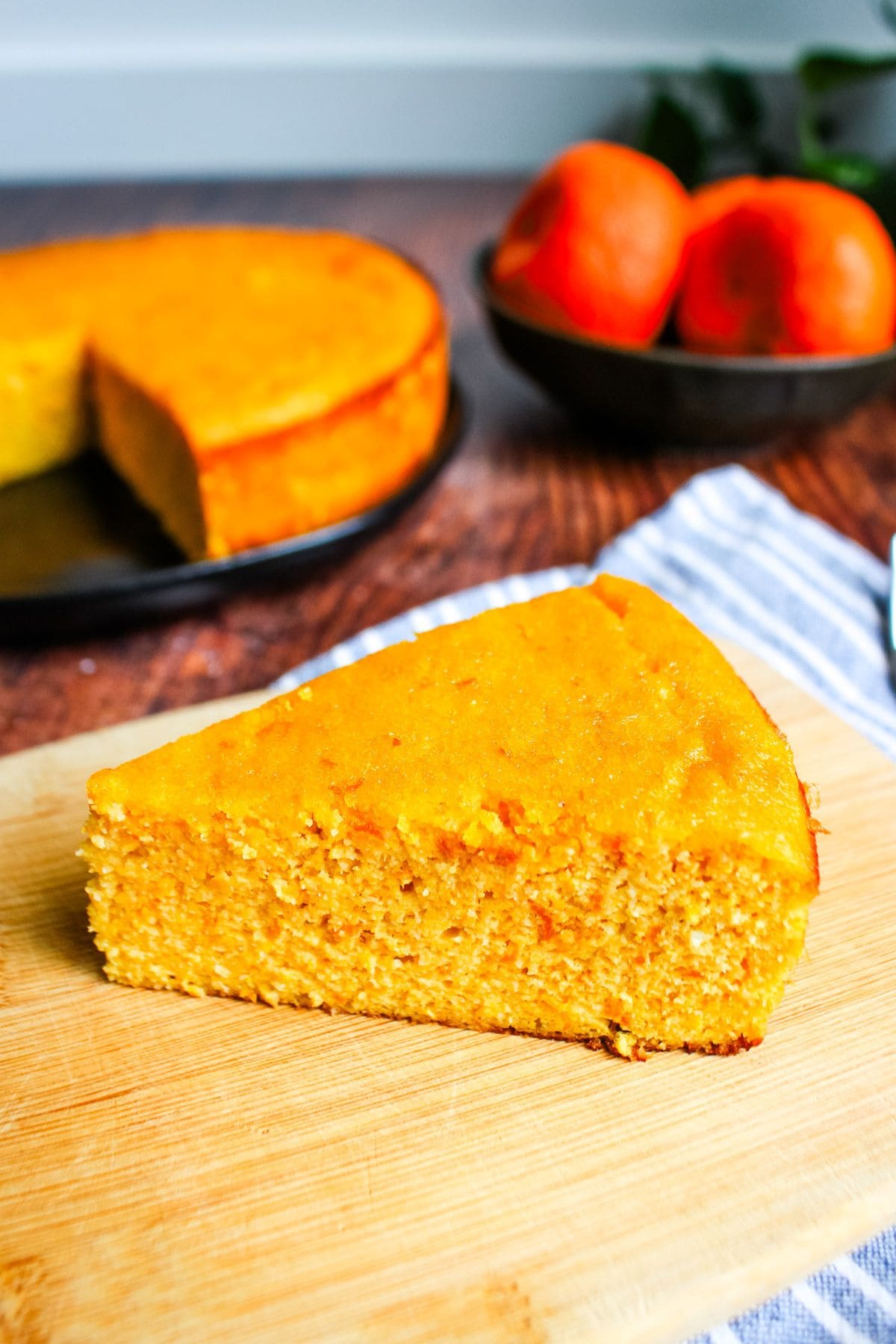 A slice of clementine cake on a wooden cutting board with the whole cake and a bowl of clementines in the back.